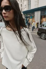 Blusa de crepe con cuello barco y manga larga. Presenta un diseño minimalista en color blanco con detalles de vivos en color negro que recorren el cuello, las mangas y los puños. Los puños incluyen botones decorativos.