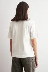 Remera color crudo de manga corta, con cuello redondo y un aplique bordado frontal con diseño floral enmarcado por pespuntes.