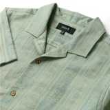 Camisa de manga corta color verde con cuello cubano y cierre frontal con botones. Presenta un diseño estampado a rayas verticales con motivos sutiles.
