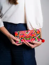 Billetera de mano con cierre perimetral, confeccionada en tela de base beige con bordado floral multicolor y vibrante en tonos fucsia, rojo y verde, con tirador de cuentas de madera y pompón rojo.