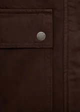 Chaqueta corta de algodón en color marrón oscuro, con cuello de efecto piel en tono contrastante. Presenta cierre frontal con cremallera y botones a presión, además de dos bolsillos frontales con solapa y cierre de botón.