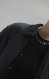 Campera tipo sobrecamisa de diseño oversize, confeccionada en microfibra opaca con patrón matelaseado geométrico. Presenta cuello, puños, cartera central y ruedo en gabardina de algodón, con cierre frontal mediante broches a presión. El diseño del ruedo simula una prenda superpuesta.