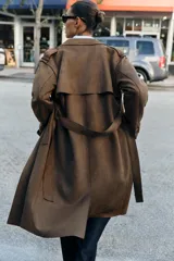Trench largo de ante sintético color marrón oscuro, de corte cruzado con doble botonadura, solapas amplias y cinturón ajustable del mismo tejido. Cuenta con bolsillos laterales de vivo, mangas largas con trabillas en los puños y detalles en los hombros.