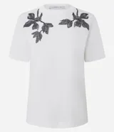 Remera blanca de manga corta y cuello redondo, confeccionada en algodón con bordados florales en tono oscuro aplicados en los hombros.