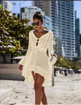 Vestido de playa corto en tejido calado color crema, con mangas largas acampanadas y diseño asimétrico con volado en el ruedo. Presenta un escote redondo con abertura frontal y cordón ajustable.