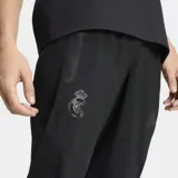 Pantalón deportivo Adidas del Real Madrid, modelo Tiro Travel, color negro. Presenta un diseño técnico con corte clásico, cintura elástica, costuras ergonómicas para mayor movilidad y bolsillos laterales. Incluye el escudo del club y el logo de la marca en tono sobre tono.