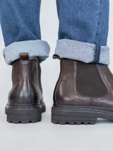 Botín Chelsea para hombre, color marrón oscuro con acabado lustroso, confeccionado en cuero. Presenta paneles elásticos laterales y suela de goma gruesa y dentada.