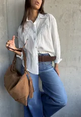 Camisa blanca de corte clásico con sutiles rayas verticales y delicado bordado floral en la parte superior delantera. Presenta cuello camisero, cierre frontal con botones y mangas largas.