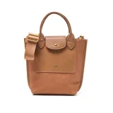 Cartera tipo tote bag de material símil cuero color habano, con cuerpo principal de tela texturada en tono similar. Posee doble asa corta, correa larga desmontable y regulable, cierre superior metálico y bolsillo interno con cierre. Presenta un bolsillo frontal con solapa de cuero y herrajes dorados.