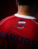 Camiseta de fútbol Umbro del Club Nacional de Football, modelo Away 2026. Diseño en color rojo con detalles en azul y blanco en el cuello y mangas. Incluye logo de Umbro, escudo del club y sponsors estampados en el frente y mangas.