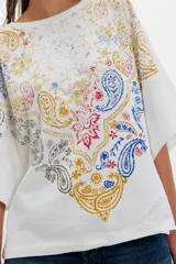 Remera de algodón blanca de corte oversize y silueta recta, con escote redondo, hombros caídos y mangas cortas amplias. Presenta una estampa frontal con diseño paisley en tonos rojo, azul, amarillo y gris.