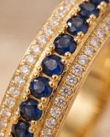 Anillo de bronce con baño de oro, diseño de banda ancha con una hilera central de circonias azules y hileras laterales de circonias transparentes.