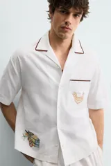 Camisa de corte relajado confeccionada en algodón, con cuello bowling y manga corta. Presenta un bolsillo en el pecho con vivo a contraste y bordados decorativos en el frente. Cierre frontal mediante botones.