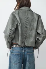 Campera biker color verde grisáceo con efecto desgastado, cuello con solapa y cierre lateral. Presenta herrajes plateados y detalles de cordones entrelazados en los codos y en la espalda.