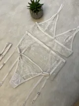 Conjunto de lencería de encaje blanco compuesto por un bralette triangular, una tanga con tiras y un portaligas integrado. El diseño presenta detalles de encaje floral y breteles regulables.