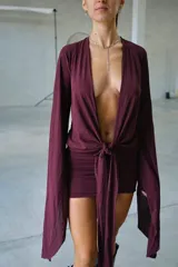 Vestido corto color bordó, con escote profundo en V, mangas largas acampanadas y detalle de nudo frontal con tiras largas. Incluye una falda ajustada tipo mini.