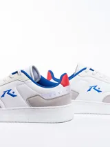 Championes urbanos blancos con detalles en gris, azul y rojo. Presentan el logo de la marca en el lateral y cordones blancos.