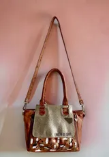 Cartera de cuero color marrón terra y dorado, con solapa de cuero texturizado, arte pintado a mano en la base, doble asa de hombro y herrajes metálicos.