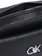 Cartera cruzada rectangular de Calvin Klein, color negro, con textura granulada en el exterior. Presenta un bolsillo frontal con cierre giratorio y el monograma metálico 'CK'. La correa es ajustable y combina una sección de cuero sintético negro con una cadena metálica plateada.