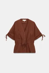 Chaqueta estilo safari de color marrón, con escote en pico, manga corta con cordones ajustables con abalorios y bolsillos delanteros de plastrón. Cuenta con cordón ajustable en la cintura y cierre frontal con botones.