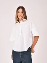 Blusa blanca de manga corta, con cuello camisero y abertura frontal parcial con botones. Presenta un corte holgado y bajo redondeado.