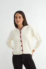 Cardigan de punto color crema con cuello redondo y cierre frontal de botones en color bordó. Presenta un detalle de costura decorativa en contraste a lo largo de la botonadura y el cuello, con mangas largas y puños acanalados.