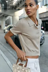 Chaqueta de manga corta y punto en color beige, con cuello polo y cierre frontal de botones. Presenta detalles de puntilla a contraste en la botonera y los puños.