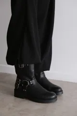 Bota de caña media en eco cuero negro, con diseño estilo biker que incluye correas con hebillas metálicas y herrajes plateados en el tobillo. Cuenta con suela plana y punta redondeada.