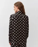 Camisa de corte recto y silueta fluida, color negro con estampado de lunares blancos. Presenta cuello clásico, cierre frontal de botones y canesú curvo sobre el pecho. Las mangas son largas, ligeramente abullonadas, con puños anchos abotonados y bajo redondeado.