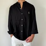 Camisa de lino blanca con cuello mao y cierre frontal de botones. Presenta un pequeño bordado oscuro en el pecho y mangas largas con puños doblados.