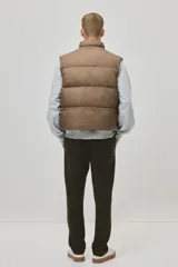 Chaleco acolchado color tostado, de nylon guateado, repelente al agua y rompevientos. Cuenta con cuello elevado, cierre bidireccional con protección para el mentón, bolsillos laterales con cierre y un bolsillo interior con botón a presión. El bajo es ajustable con cordón elástico y topes.