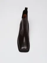 Botas tipo chelsea de cuero color marrón oscuro con acabado brillante. Presentan punta cuadrada con detalle de puntera metálica, elásticos laterales y taco cuadrado bajo.