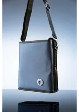 Bolso tipo bandolera color marrón, de diseño rectangular y minimalista. Cuenta con una correa larga ajustable con el nombre de la marca grabado y un aplique circular con el logo en la parte inferior frontal.