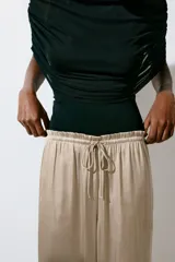 Pantalón fluido de satén color beige, de corte holgado y piernas amplias, con cintura elástica fruncida y cordón ajustable.