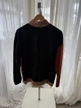 Buzo de corderito con diseño tricolor, que combina cuerpo en tono tostado, una manga negra y otra en color ladrillo. Cuenta con cuello alto con botones frontales y bolsillo tipo canguro.