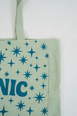 Bolso tote de tela con estampado de rayas verticales rosadas y blancas, laterales y asas en color beige. Presenta la palabra "BONJOUR" en verde en el centro y "INDIAN" en la parte inferior.