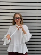 Blusa blanca de textura arrugada, con escote en V y lazos para anudar. Presenta mangas largas con puños acampanados y un diseño con volado en el ruedo.