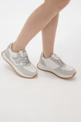 Zapatilla deportiva de diseño urbano, combinando paneles de tela blanca y gamuza sintética en tono gris claro. Cuenta con cordones grises, suela de plataforma con entresuela blanca y base dentada en color caramelo.