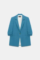 Blazer azul con cuello solapa, hombreras y mangas por debajo del codo con detalle drapeado. Presenta falsos bolsillos de vivo en el delantero, forro interior y cierre frontal con un botón.