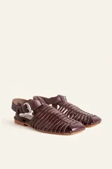 Sandalia de cuero negro estilo huarache, con puntera cerrada mediante tiras finas entretejidas a mano, tira ajustable al tobillo con hebilla metálica y suela plana con taco de 1 cm.