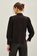 Camisa negra de algodón con detalles de encaje calado en el pecho, hombros y puños. Presenta cuello alto con terminación de encaje, cierre frontal con botones y mangas largas abullonadas.