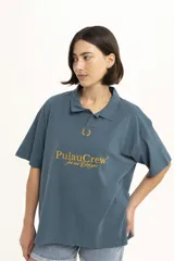 Remera de manga corta color verde oscuro, estilo polo con cuello y abertura frontal. Presenta un bordado central con el texto "PulauCrew" y un laurel en la parte superior.
