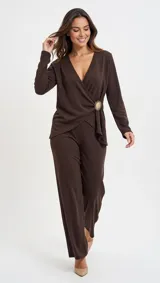 Blusa de color marrón con acabado brillante, diseño cruzado tipo sobre con escote en V y manga larga. Presenta un detalle de broche decorativo en forma de sol en el lateral de la cintura.