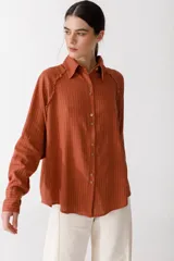 Camisa color ladrillo con rayas verticales finas color crema, de corte recto y mangas largas con puños abotonados. Presenta cuello camisero, cierre frontal con botones y detalle de flecos en los hombros.