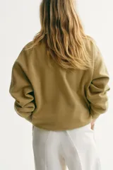 Buzo oversized color beige claro, con cuello redondo, hombros caídos y el texto "ATELIER DE MODE" en relieve con textura de felpa blanca en el frente. Cuenta con puños, cuello y dobladillo en ribete acanalado.