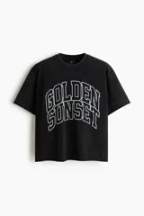 Remera boxy de algodón negro con efecto deslavado, corte oversized y mangas cortas. Presenta un estampado gráfico frontal con la inscripción "Golden Sunset" en estilo universitario.