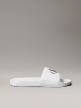 Ojotas tipo slide Calvin Klein Jeans, color blanco, con banda superior de lona de algodón que presenta el monograma de la marca en color negro. Posee suela de EVA con plantilla ergonómica moldeada.