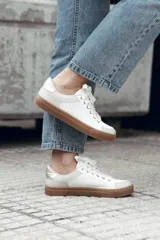 Zapatilla de estilo urbano en tonos marfil, con detalles en color dorado en el talón y ojales metálicos. Presenta suela de goma color caramelo y cordones al tono.