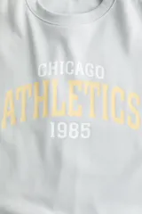 Buzo oversize celeste de cuello redondo, con hombros caídos, puños y bajo acanalados, y diseño de texto frontal con la inscripción "CHICAGO ATHLETICS 1985".