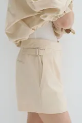 Short de mezcla de algodón y lino color beige, con pinzas delanteras, raya marcada al frente y ajuste con hebillas metálicas en los laterales.